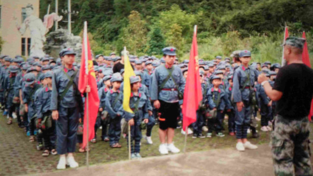 붉은 군대 유니폼을 입은 학생들이 마오 주석의 초상화와 “인민을 위하여 봉사(為人民服務)”라는 구호가 인쇄되어 있는 배낭을 옮기고 있다.